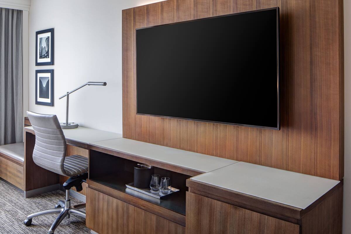 A modern hotel room with a large flat-screen TV mounted on the wall, a wooden desk with a chair, and a small shelf with glasses.