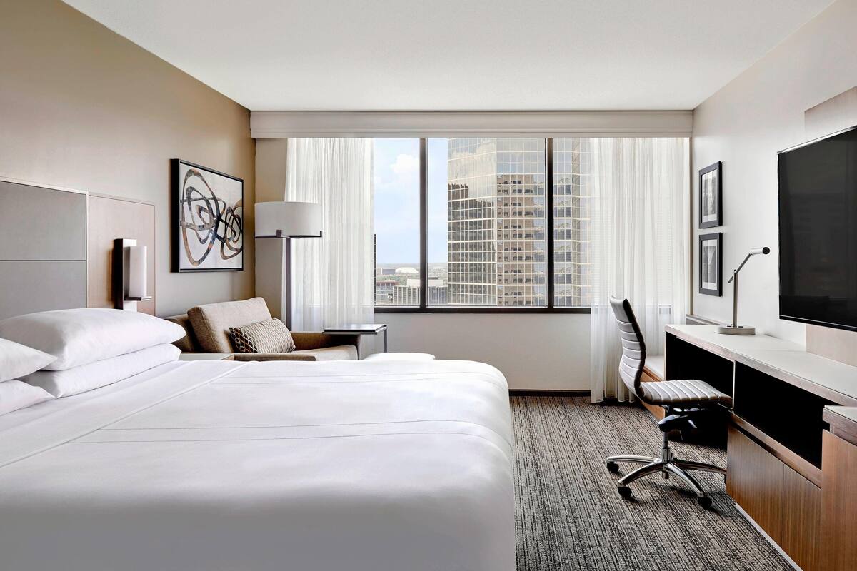 A hotel room with a large bed, a desk, a chair, a TV, and a window with a city view.