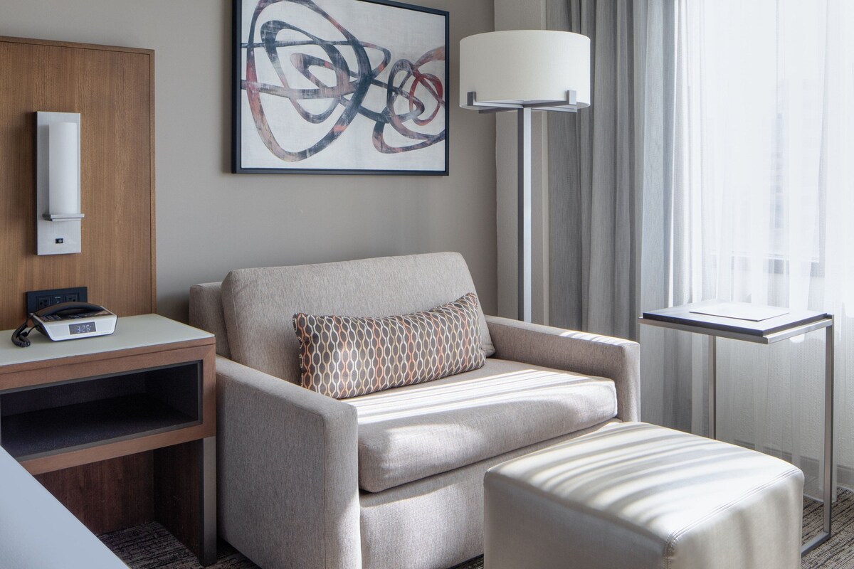 A modern hotel room with a sofa, a small table with a lamp, a painting on the wall, and a window with curtains.
