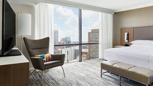 A hotel room with a large bed, a desk with a television, a chair with a colorful cushion, a bench, and a city view through large windows. - Houston Marriott Medical Center/Museum District (Houston)
