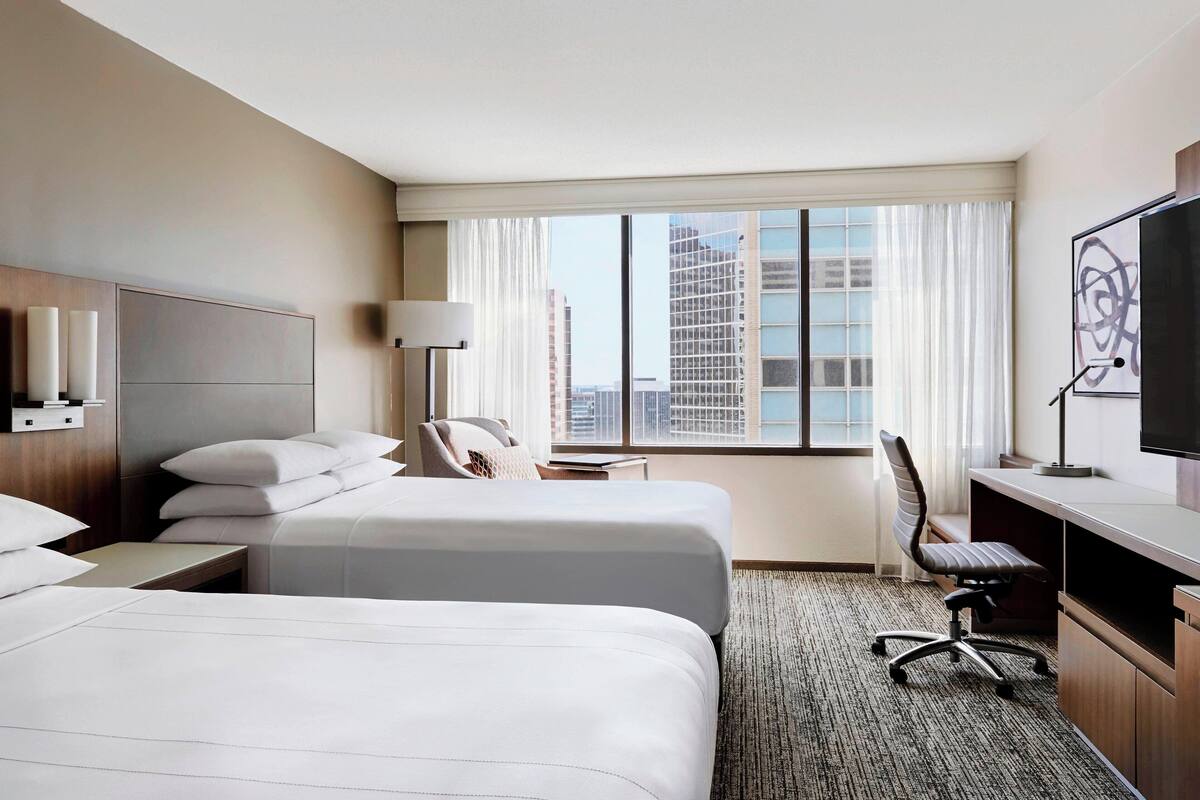 A hotel room with two beds, a desk, a chair, a TV, and a window with a city view.