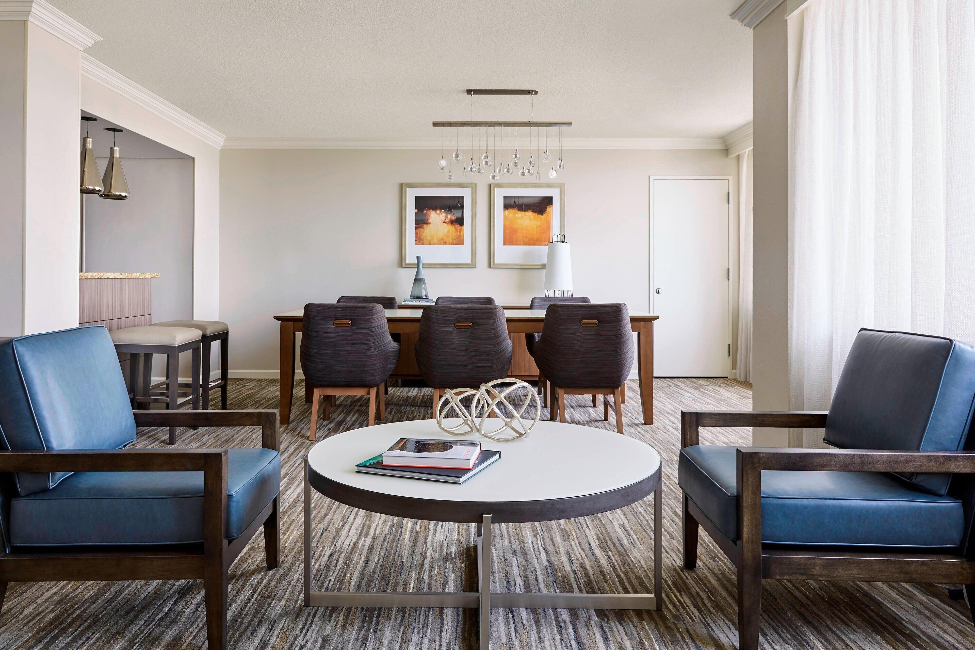 Una habitación de hotel moderna con una mesa redonda de comedor, cuatro sillas, una estantería y un amplio ventanal con cortinas.