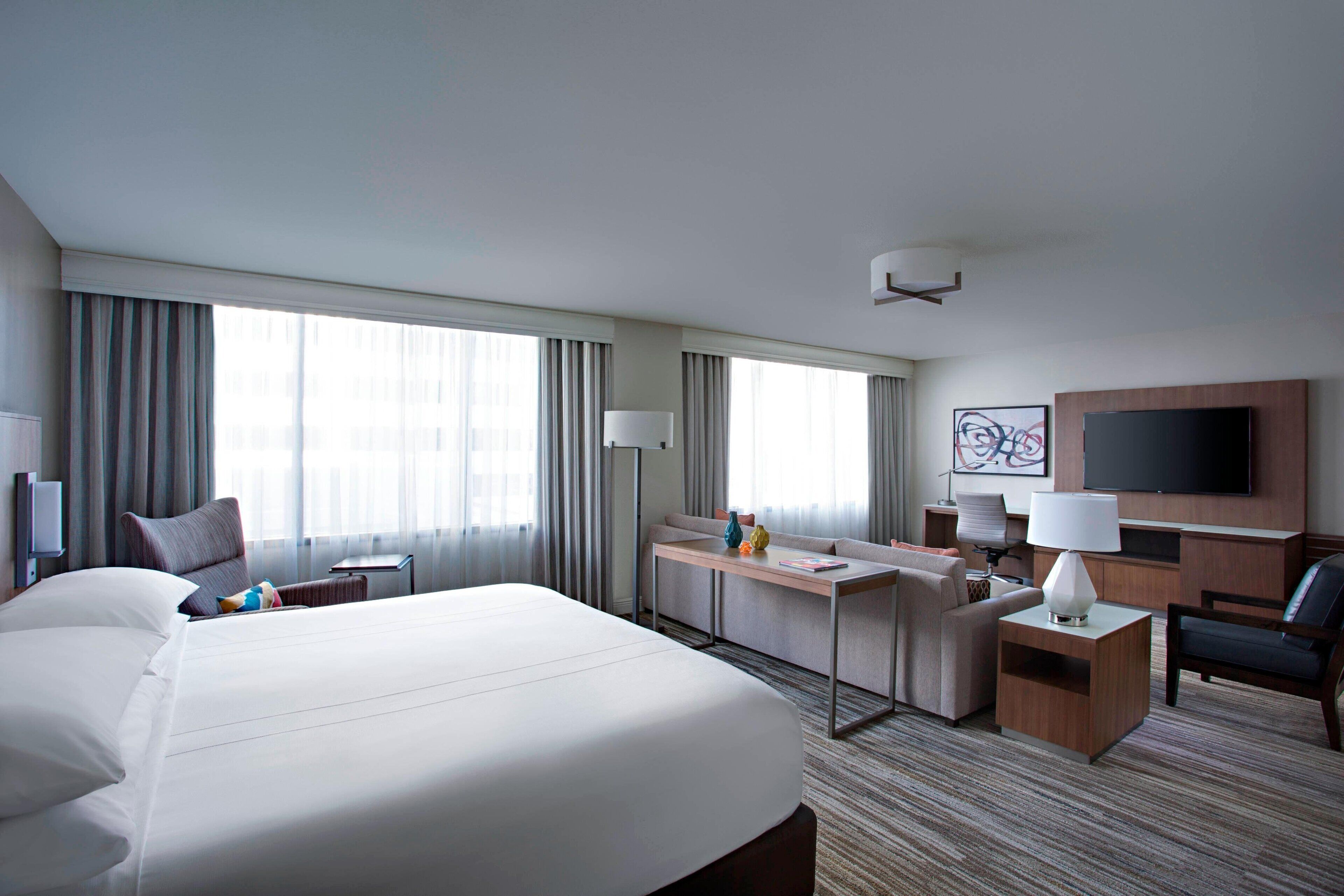 Quarto de hotel com cama grande, escrivaninha, cadeira, televisão e janela com cortinas.