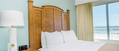 A hotel room with a large bed, a wooden headboard, a nightstand with a lamp, and a view of the beach through the window.