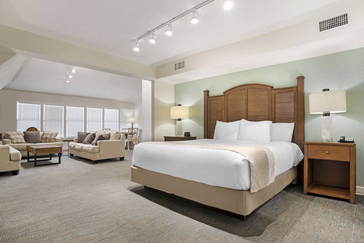 A spacious hotel room with a large bed, two bedside tables with lamps, a sofa set, and a view of the outdoors through large windows.