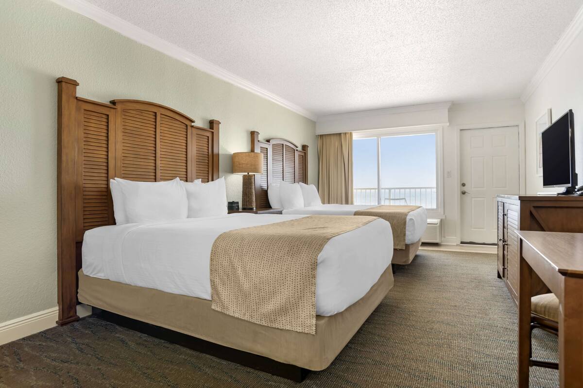 A hotel room with two beds, a TV, a desk, and a balcony with a view.