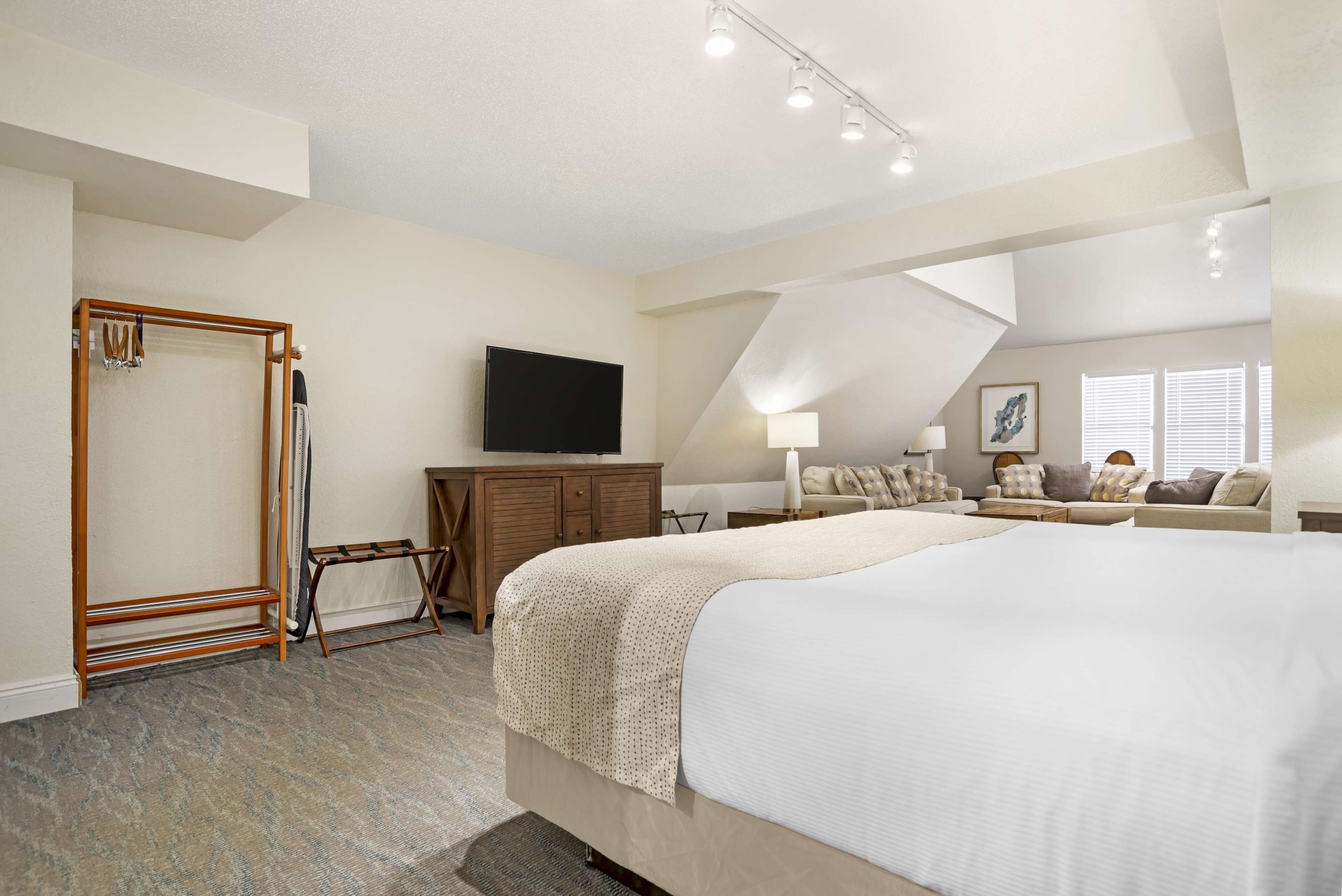 A spacious bedroom with a large bed, a TV mounted on the wall, a wooden wardrobe, and a seating area with a sofa and armchairs.