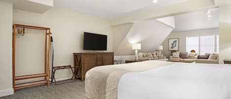 A spacious bedroom with a large bed, a TV mounted on the wall, a wooden wardrobe, and a seating area with a sofa and armchairs.