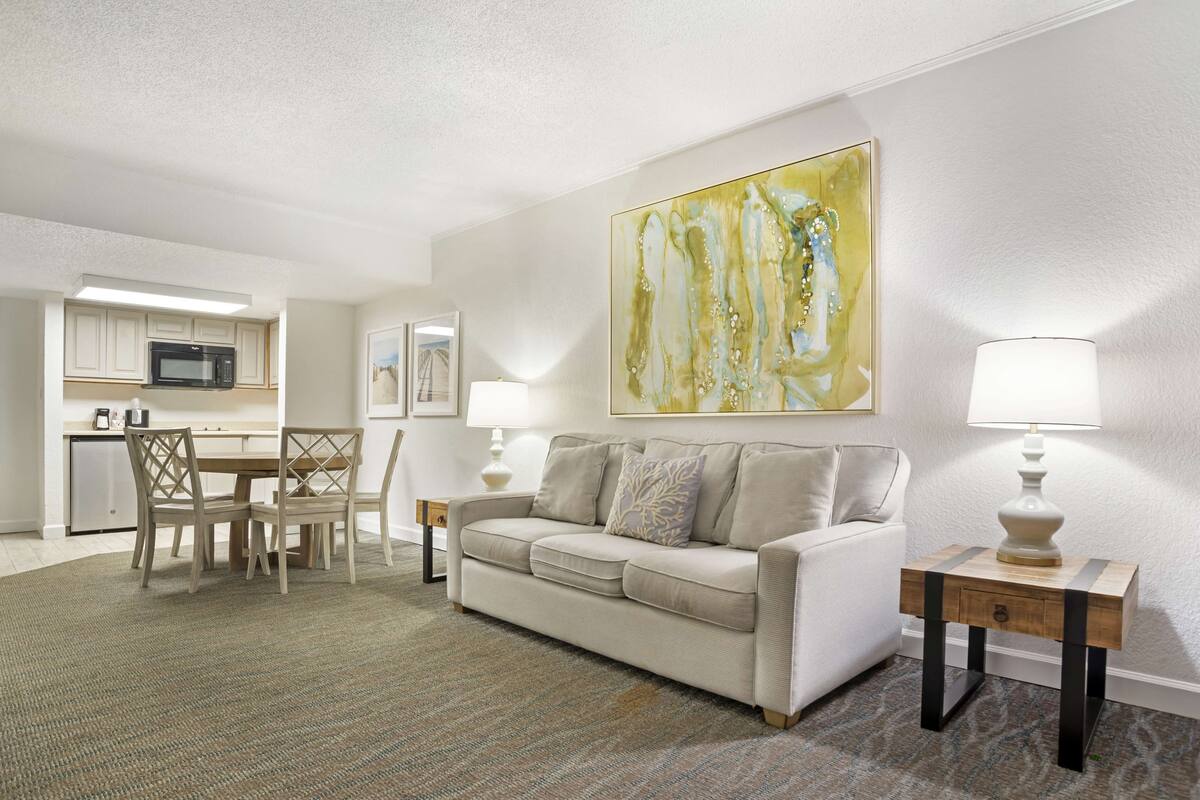 A living room with a sofa, a coffee table, and a dining area with chairs and a table. There is a kitchen with a microwave and cabinets. A painting is on the wall, and there are two lamps on side tables.