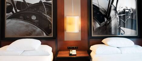 A hotel room with two beds, a nightstand with a lamp, and two framed black and white photographs of boats.