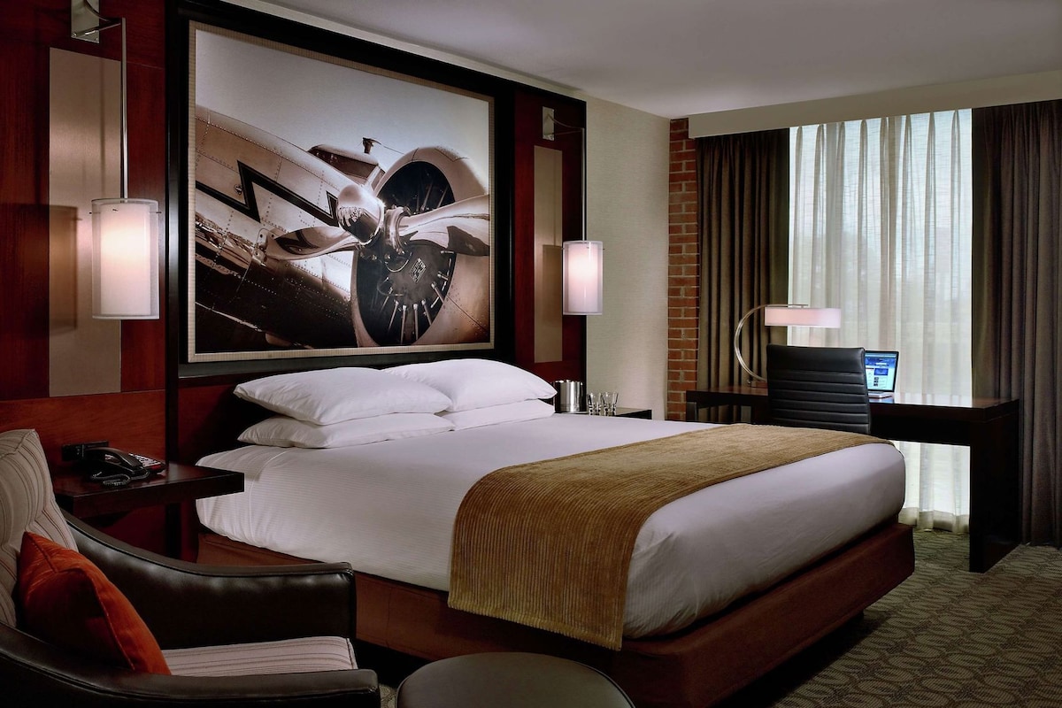 Hotelkamer met een groot bed, een bureau met een computer, een stoel, een lamp en een groot abstract schilderij aan de muur.