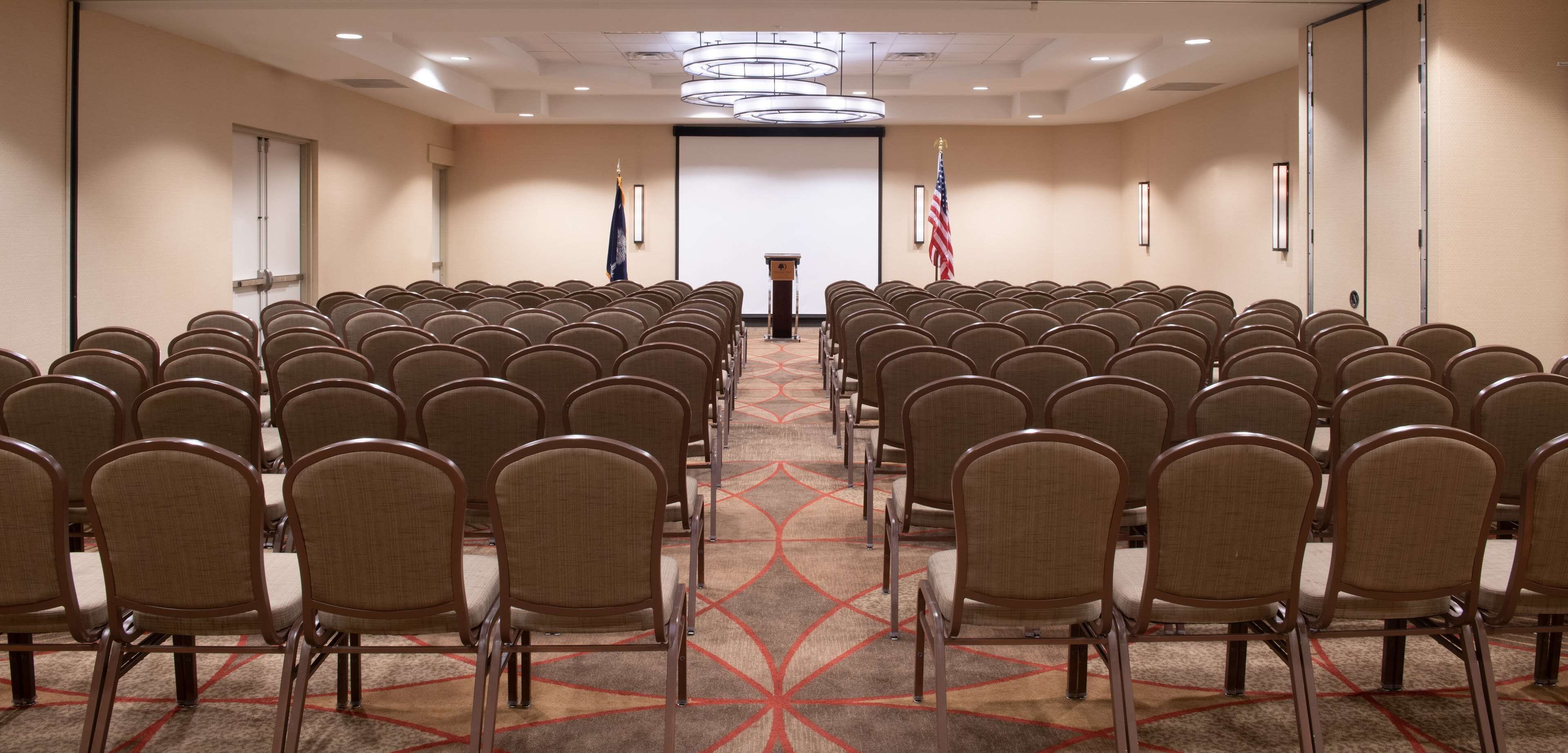 Une salle de conférence avec des rangées de chaises disposées face à un pupitre, des drapeaux accrochés aux murs et un sol recouvert de moquette.