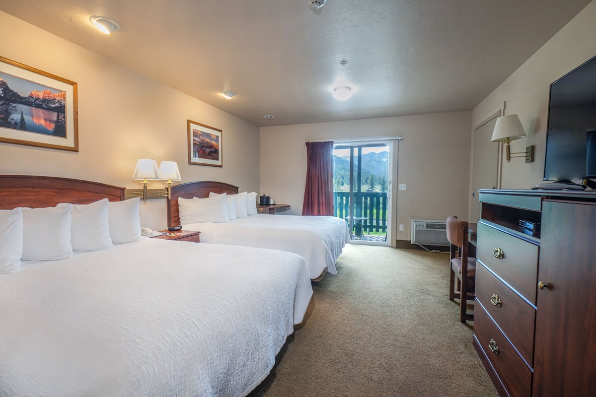 A hotel room with two beds, a dresser, a TV, a lamp, and a window with a view of mountains.