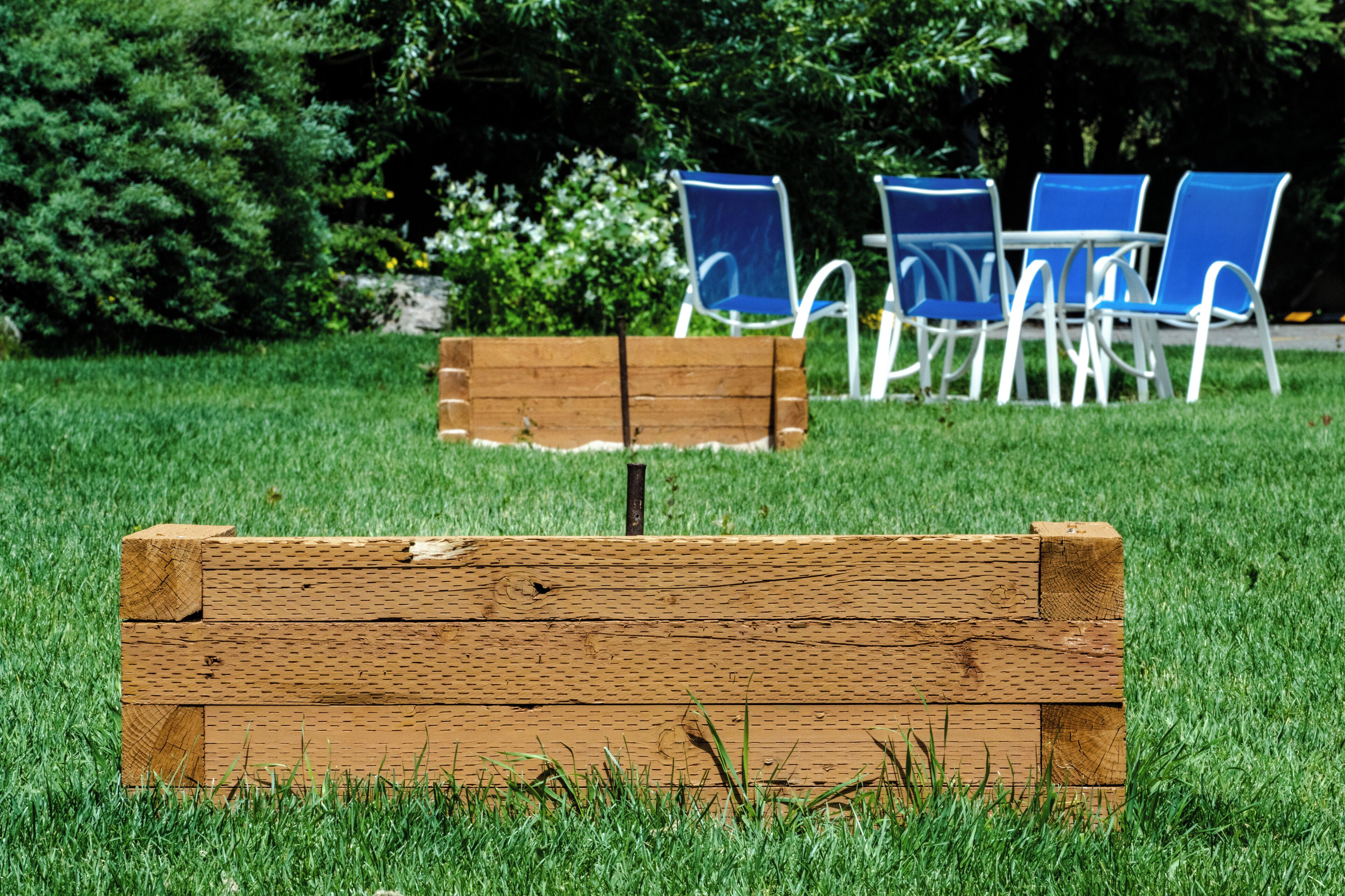 Un banc en bois se trouve dans un espace verdoyant, avec des chaises bleues et une table en arrière-plan.