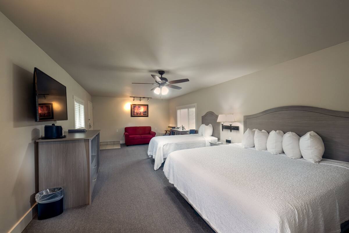 A hotel room with two beds, a desk, a red chair, a ceiling fan, and a TV mounted on the wall.
