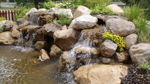 A small waterfall cascading over rocks into a pond, surrounded by lush greenery and a red fence in the background. - Flying Saddle Resort (Alpine)