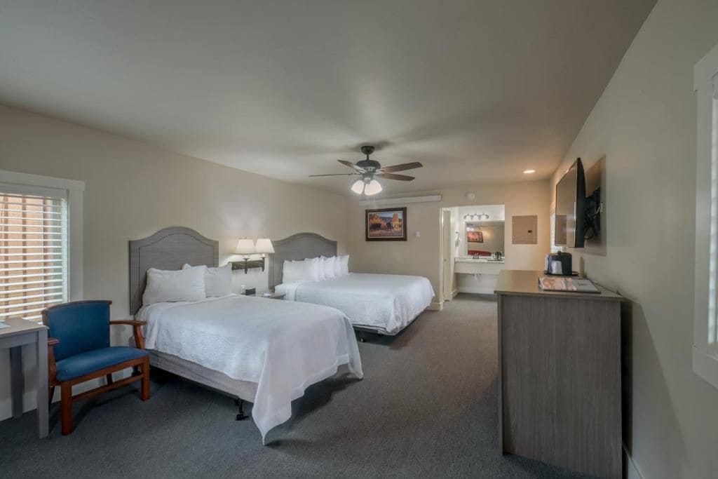 A hotel room with two beds, a desk, a chair, a desk lamp, a ceiling fan, and a television.