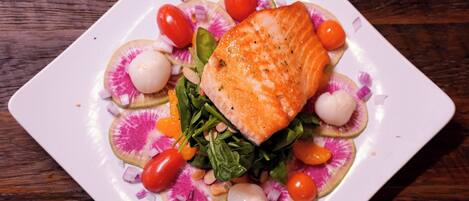 A square white plate with a piece of salmon, cherry tomatoes, radish slices, and greens on a wooden table.