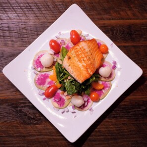 A square white plate with a piece of salmon, cherry tomatoes, radish slices, and greens on a wooden table. - Flying Saddle Resort (Alpine)