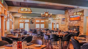 A restaurant with multiple tables and chairs, a stone wall with a fireplace, and various hanging lights. - Flying Saddle Resort (Alpine)