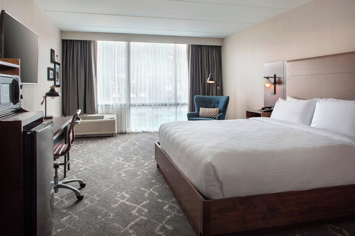 A hotel room with a large bed, a desk with a computer, a chair, a desk lamp, and a large window with curtains.