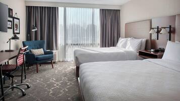 Habitación de hotel con dos camas, un escritorio con silla, una lámpara y un ventanal con cortinas.