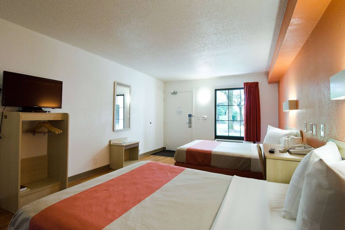 Ein Hotelzimmer mit zwei Betten, einem Fernseher, einem Schreibtisch und einem Fenster mit Vorhängen.