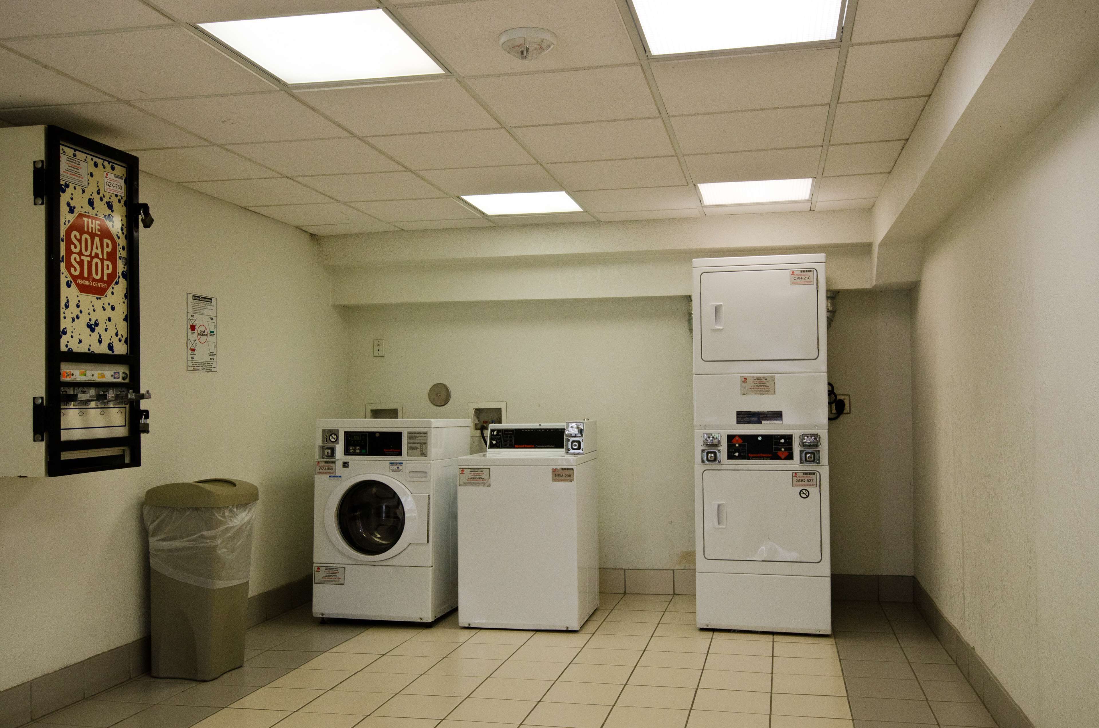 A laundry room with three washers and dryers, a trash can, and a sign on the wall.