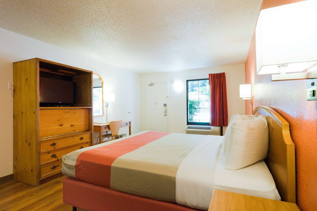 Ein Hotelzimmer mit Bett, einem Holzschrank, einem Schreibtisch mit Stuhl, einem Fenster mit Vorhängen und einer Wandlampe.