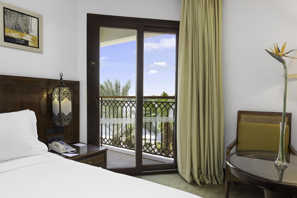 A hotel room with a bed, a desk with a phone, a chair, a balcony with a view, and a painting on the wall.