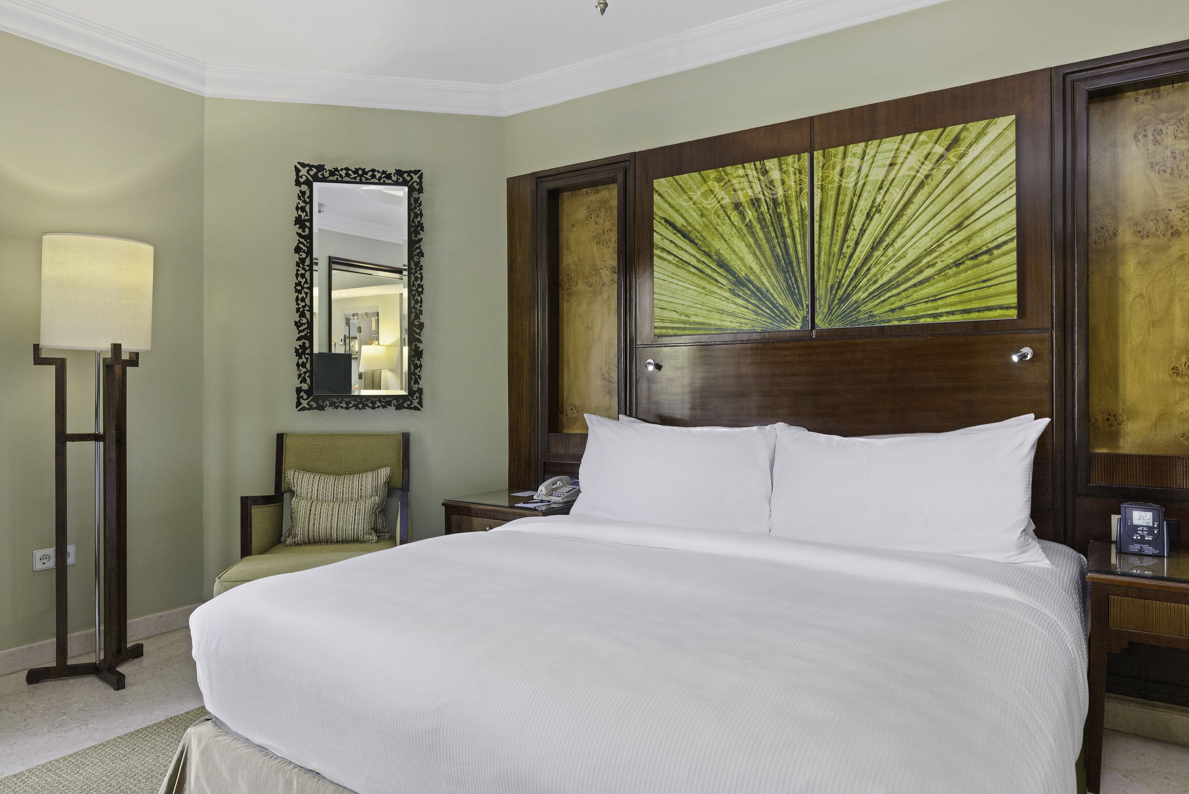A neatly made bed with white linens, a bedside table with a lamp, a chair with a cushion, and a large mirror on the wall.