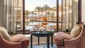 Ein Zimmer mit Glastisch, gedecktem Frühstückstisch, zwei Sesseln und Blick auf Pool und Palmen.