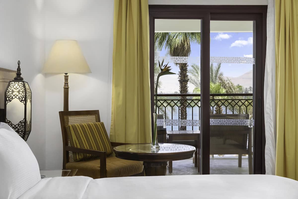 A hotel room with a bed, a chair, a table, a lamp, and a view of a balcony with palm trees and a mountain.