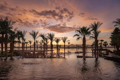 A waterfront resort with palm trees, a pool, and a sunset view.
