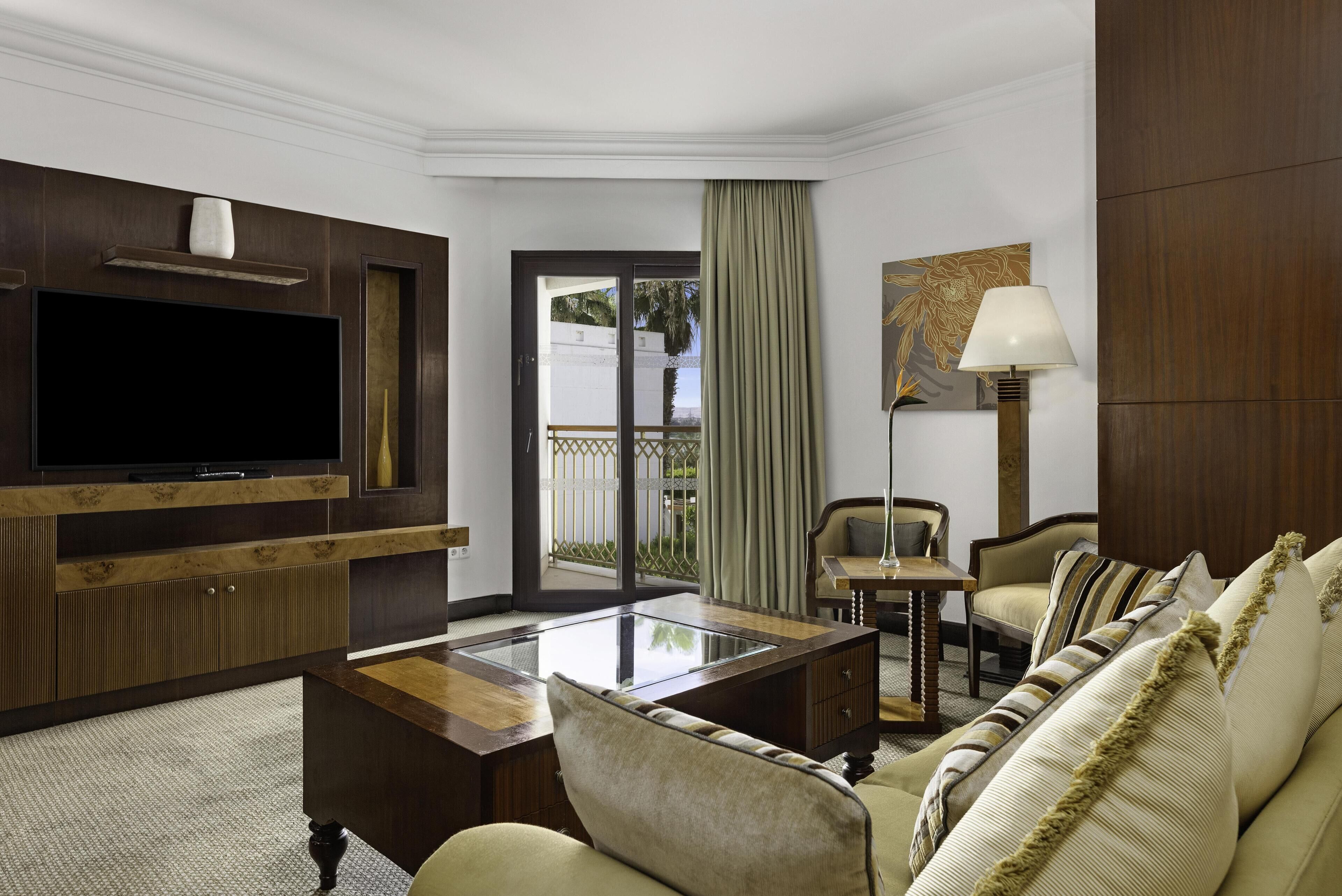 A hotel room with a flat-screen TV, a sofa, a coffee table, a lamp, and a view of the outdoors.