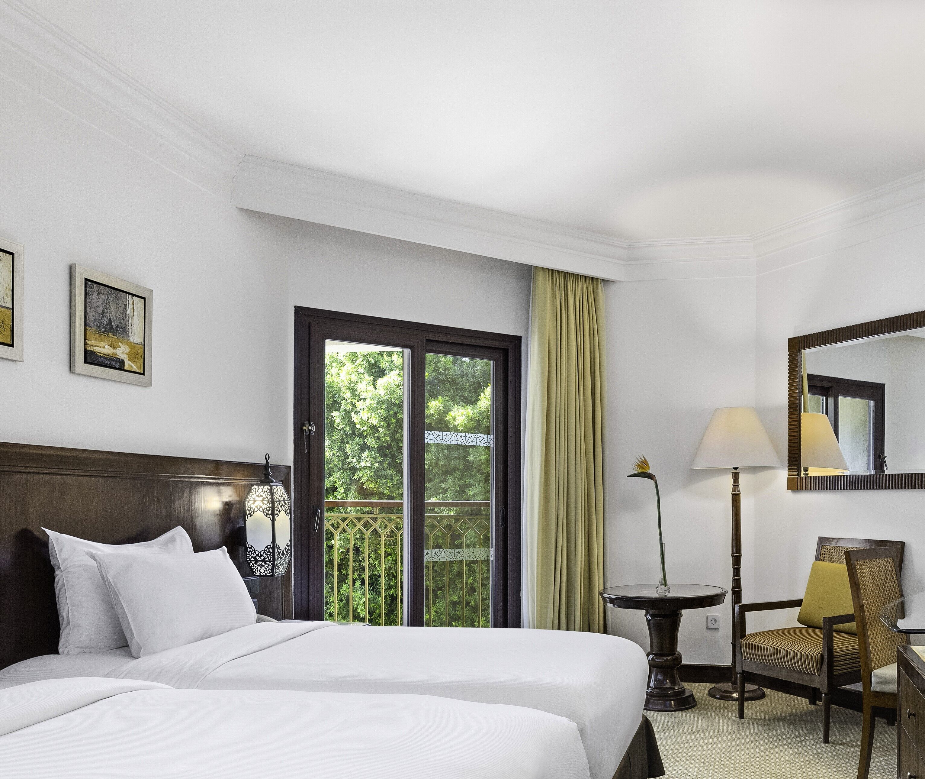 A hotel room with a large bed, a desk, a chair, a lamp, and a view of greenery through the window.