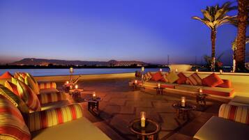 An outdoor seating area with sofas and tables, a palm tree, and a view of the water.