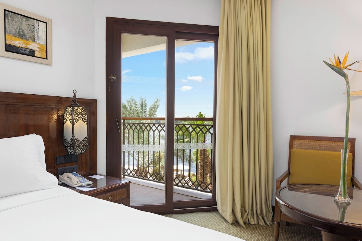 A hotel room with a bed, a desk with a telephone, a chair, a balcony with a view of palm trees, and a decorative plant.