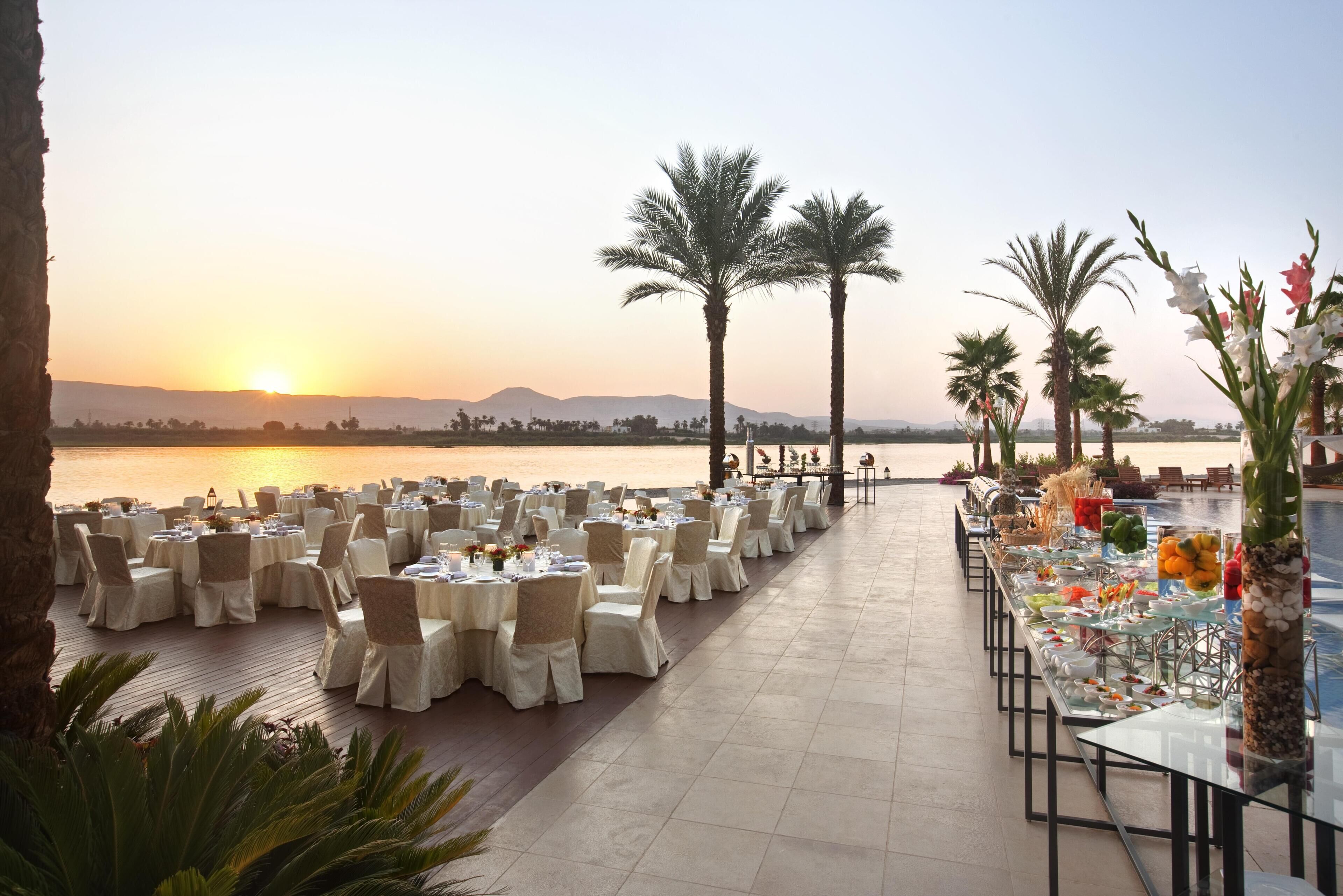 A banquet setup with tables and chairs arranged for an event, overlooking a body of water and a sunset.