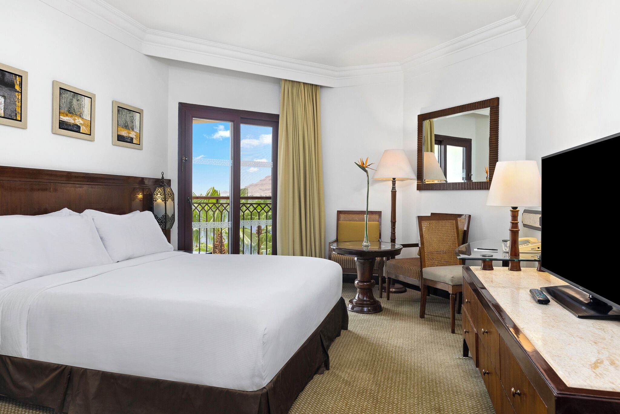 Habitación de hotel con una cama grande, un escritorio con lámpara, una silla, televisión y balcón con vistas.