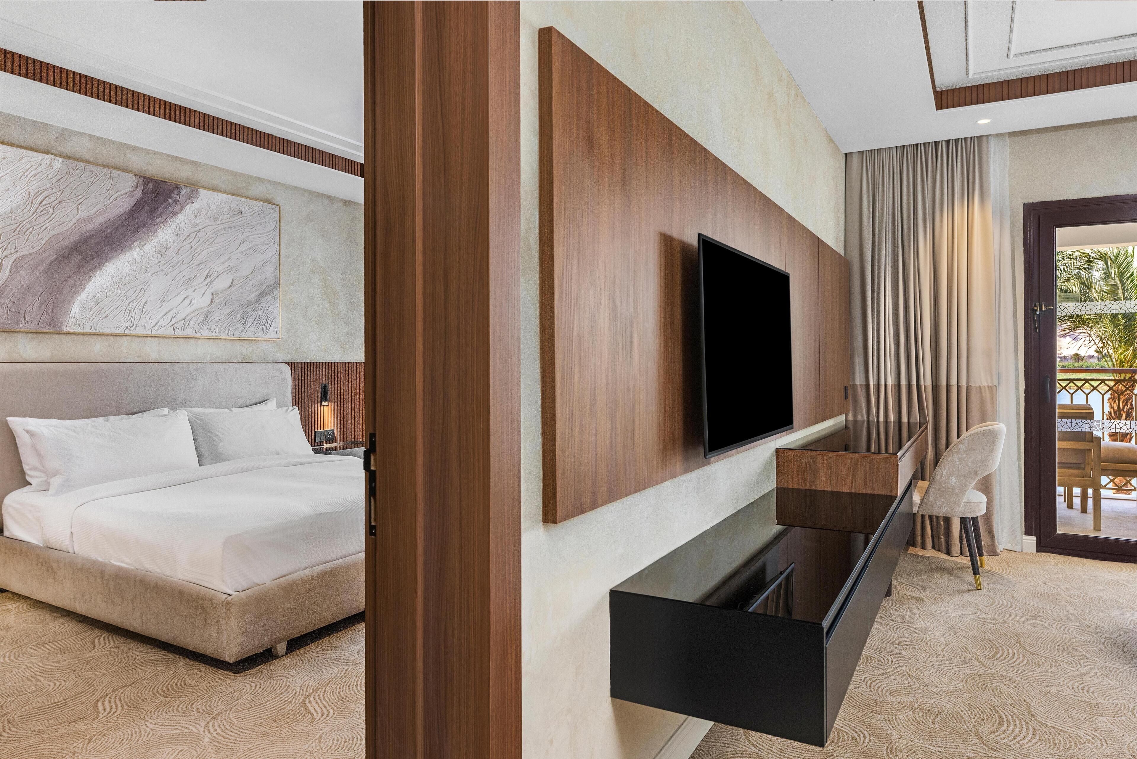 A modern hotel room with a bed, a flat-screen TV mounted on the wall, a built-in desk with a chair, and a large abstract painting above the bed.