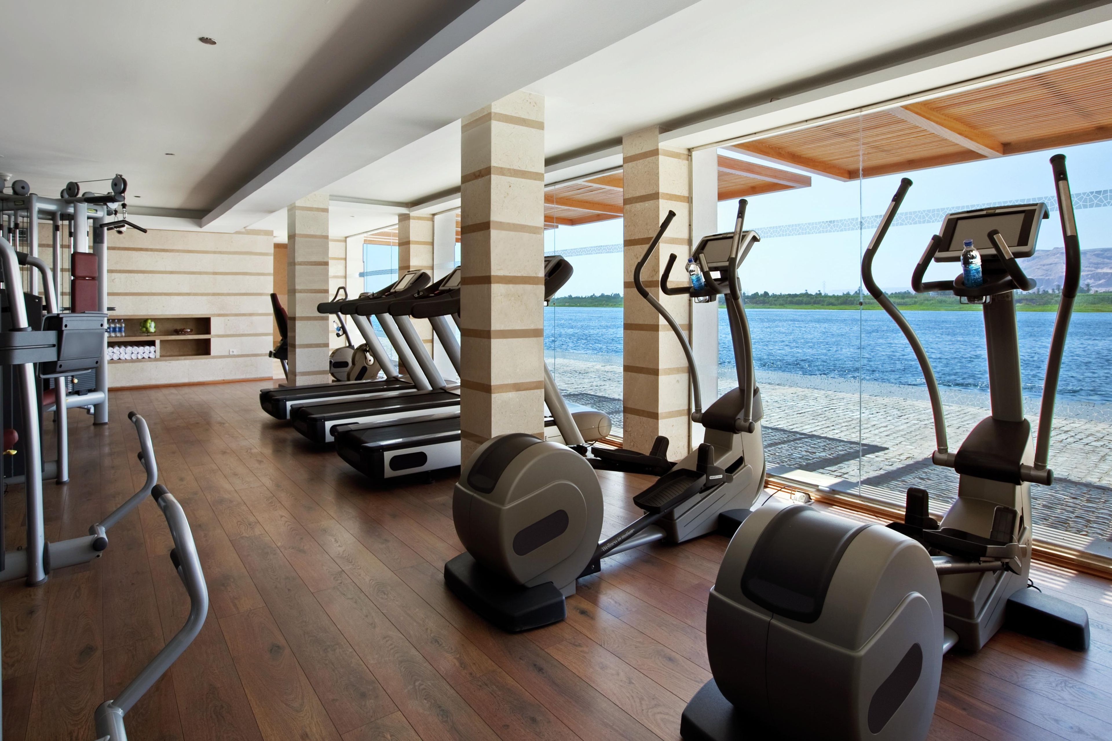 A gym with treadmills, elliptical machines, and weight equipment, with a view of a body of water.