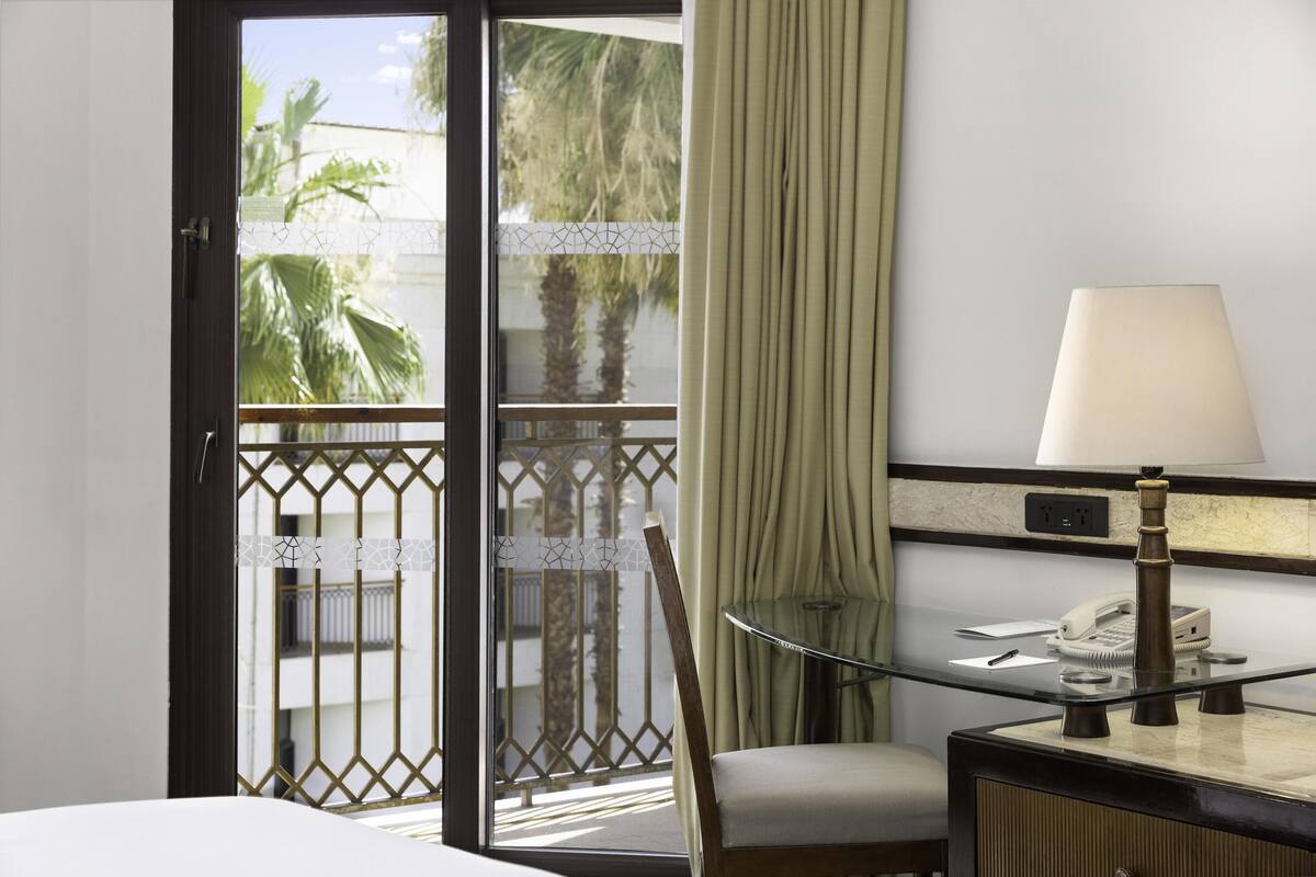 A hotel room with a bed, a desk with a lamp, a chair, and a view of palm trees through a glass door.