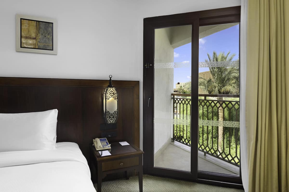 A hotel room with a bed, bedside table with a lamp, a telephone, and a view of a balcony with greenery.