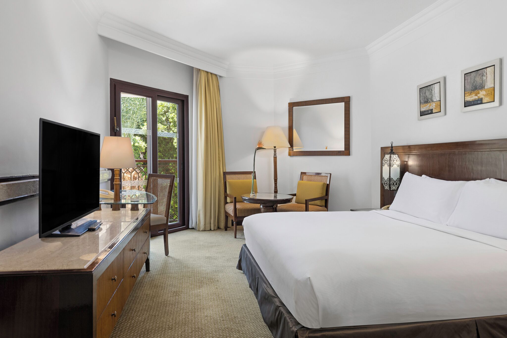 Une chambre d’hôtel comprenant un lit, une télévision, un bureau avec une lampe, une chaise, une petite table et une fenêtre avec des rideaux.