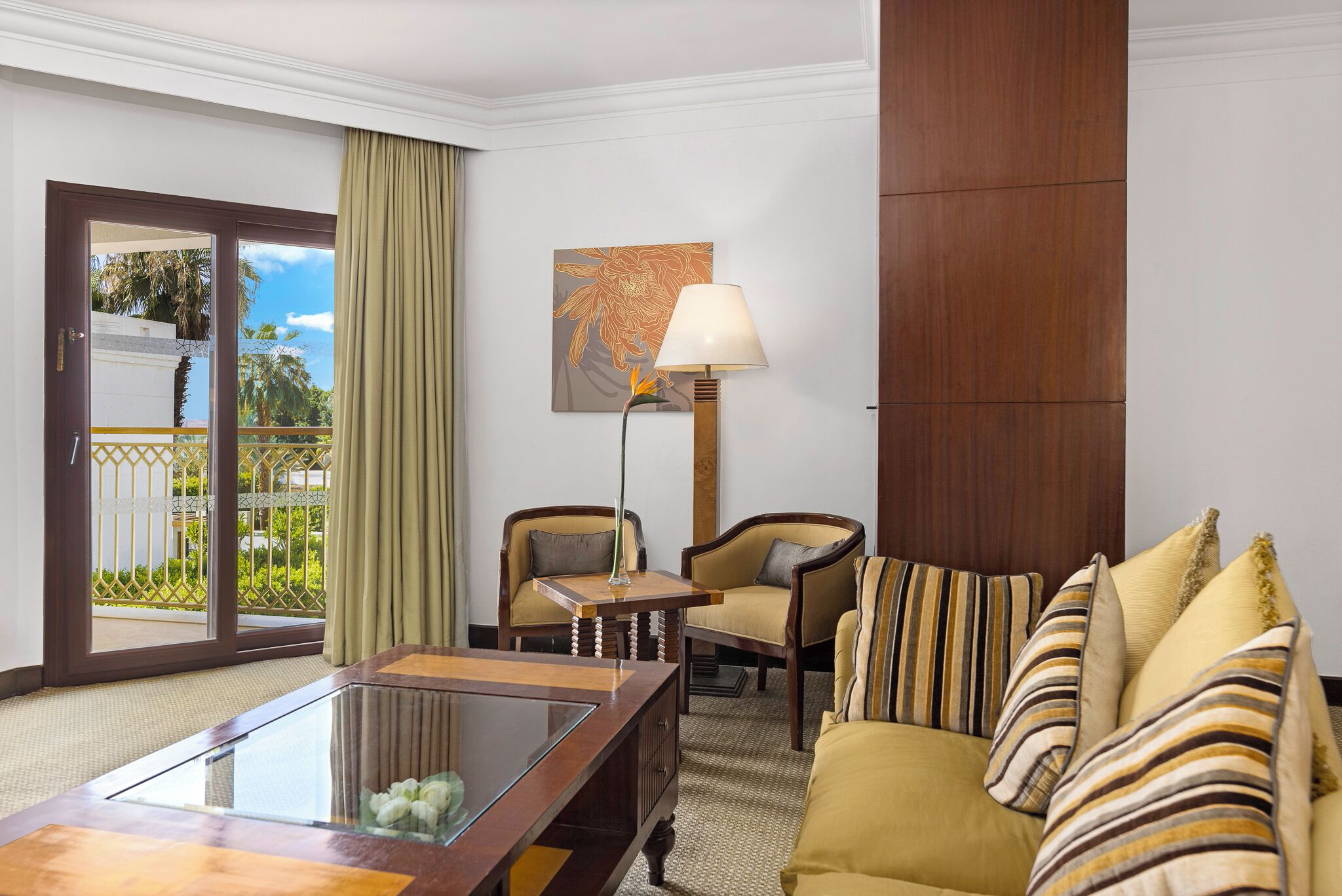 A hotel room with a glass coffee table, two armchairs, a sofa with striped cushions, a floor lamp, and a large window with a view of palm trees.