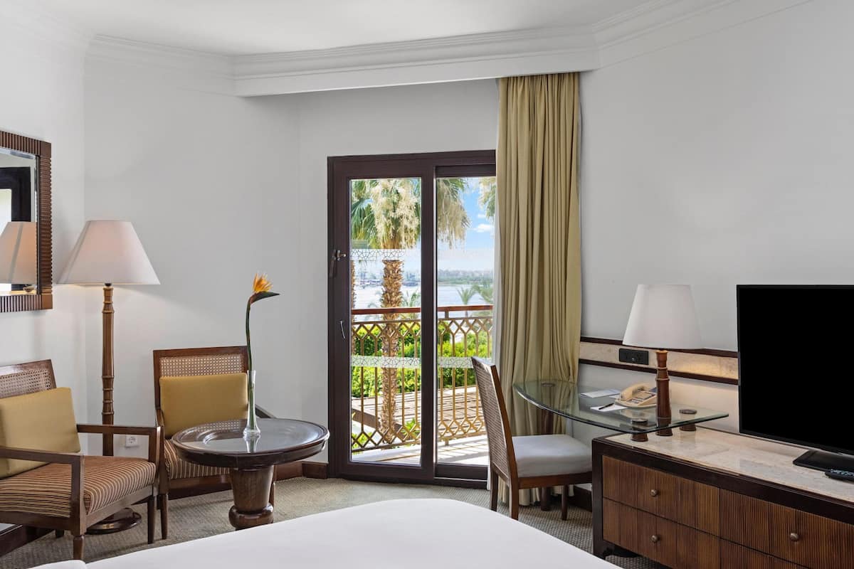 A hotel room with a bed, a desk with a television, a chair, a table, and a balcony with a view.