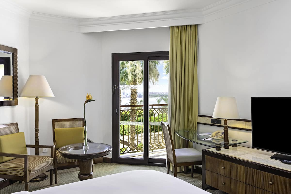 A hotel room with a bed, a desk with a lamp, a chair, a glass table, a television, and a sliding glass door leading to a balcony with a view.