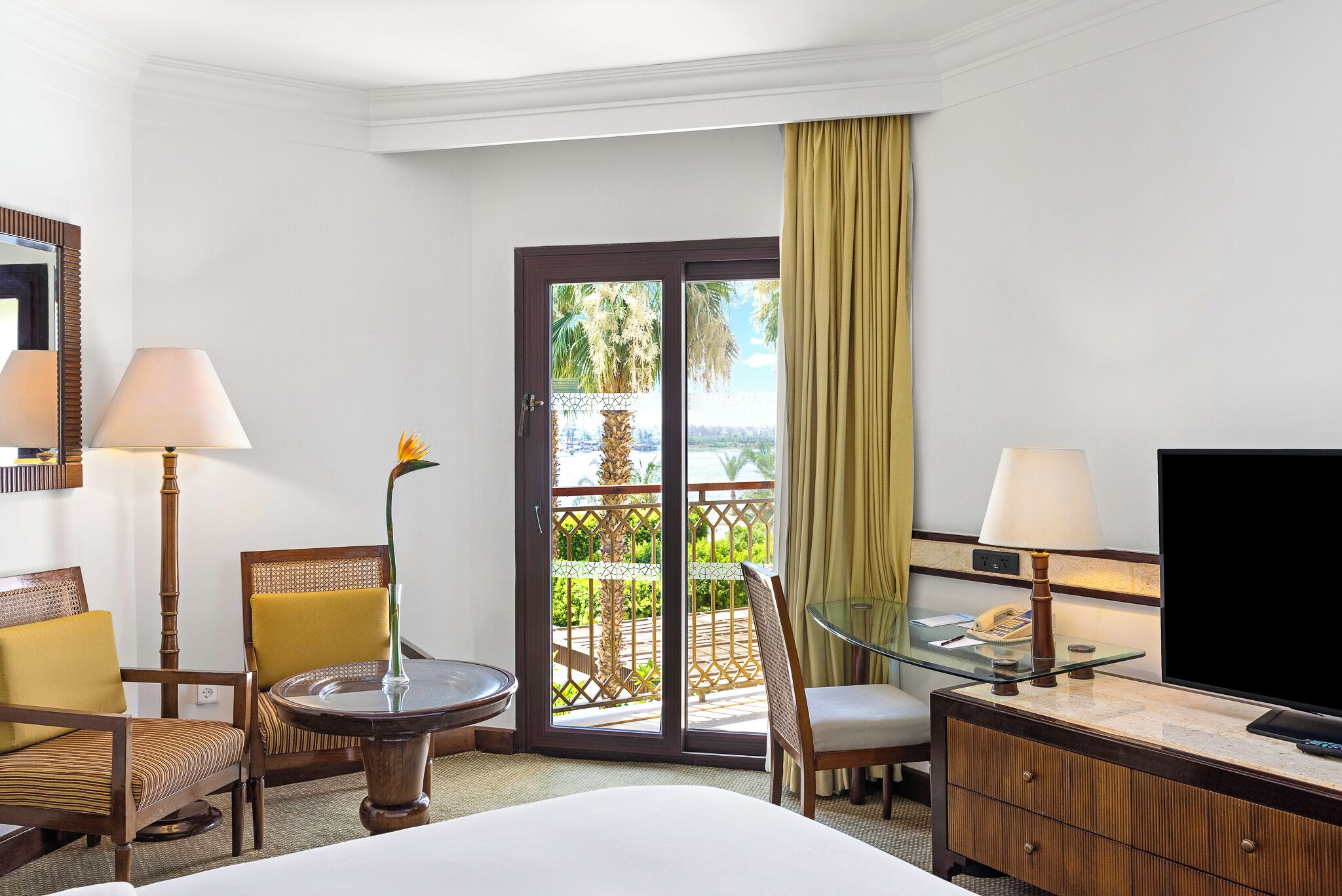 Una camera d'albergo con un letto, due poltrone, una scrivania con televisore, un tavolino in vetro e un balcone con vista.