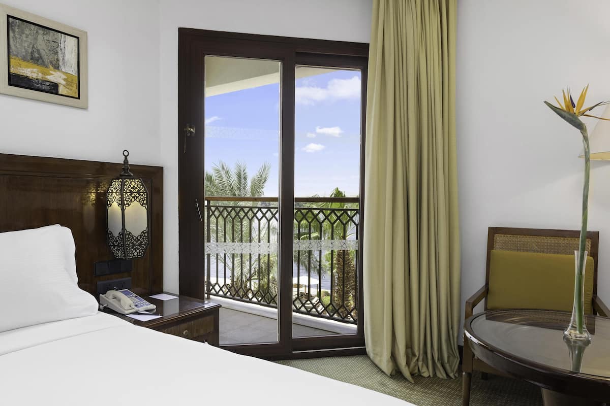 A hotel room with a bed, a desk with a phone, a chair, a balcony with a view, and a painting on the wall.